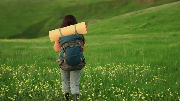 Mujer viajera libre camina a través del prado con flores en verano. Chica viajero viaja a lo largo de la pendiente de la colina verde, meditación, ecoturismo, senderismo. Una joven activa con mochila hace senderismo —  Fotos de Stock