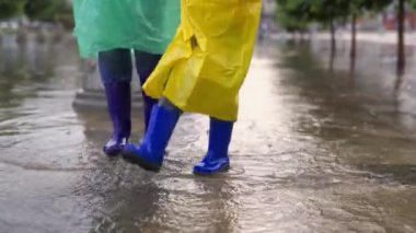 Mutlu aile lastik botlarla su birikintilerinde yürüyor. Bebeğim, annesi yağmurlu yaz parkında yağmurda yağmurda yağmurla yürür, çocuk sevinir, ayaklarının altından su akar. Çocuk su birikintisine atlıyor..