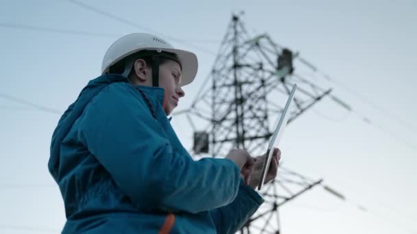 Vrouw power engineer in een witte helm controleert de stroomleiding met behulp van gegevens van elektrische sensoren op een tablet. Hoogspanningsleidingen bij zonsondergang. Distributie en levering van elektriciteit. schone energie — Stockvideo