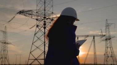 Beyaz kasklı kadın güç mühendisi tabletteki elektrik sensörlerinden elde edilen verileri kullanarak güç hattını kontrol ediyor. Gün batımında yüksek voltajlı elektrik hatları. Dağıtım ve elektrik tedariği. temiz enerji