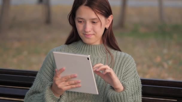 Студентка учится за планшетным компьютером на открытом воздухе. Фрилансер работает онлайн. Девушка с цифровой планшеткой печатает сообщения в парке на скамейке. Молодая женщина работает на планшете в парке на скамейке. — стоковое видео