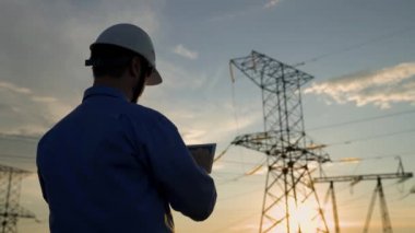 Beyaz miğferdeki güç mühendisi güç hattını tabletteki bilgisayarla kontrol ediyor, güç sisteminin uzaktan kumandası. Gün batımında yüksek voltajlı elektrik hatları. Dağıtım ve elektrik tedariği. Enerji