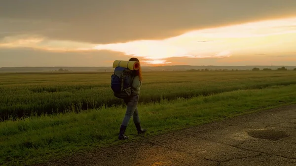 Aktivní zdravá běloška s batohem kráčí po silnici v paprscích západu slunce. Mladá žena turistika v letní přírodě. Cestovatel cestuje na venkově, meditace, ekoturistika, turistika. — Stock fotografie