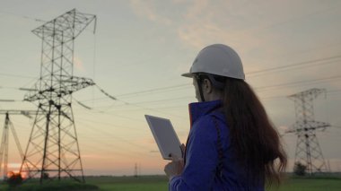 Beyaz kasklı kadın güç mühendisi tabletteki elektrik sensörlerinden elde edilen verileri kullanarak güç hattını kontrol ediyor. Gün batımında yüksek voltajlı elektrik hatları. Dağıtım ve elektrik tedariği. temiz enerji