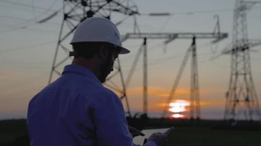 Beyaz miğferdeki güç mühendisi güç hattını tabletteki bilgisayarla kontrol ediyor, güç sisteminin uzaktan kumandası. Gün batımında yüksek voltajlı elektrik hatları. Dağıtım ve elektrik tedariği. Enerji