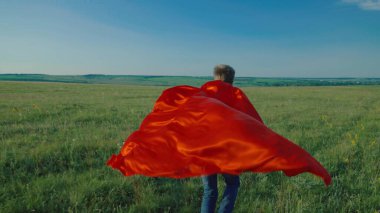 Mutlu bir çocuk, süper kahramanı oynayan bir çocuk, kırmızı pelerinli, rüzgarda çırpınan bir pelerinle yeşil bir alanda koşuyor. Çocuk oyunları ve hayalleri. Ağır çekim. Çocuk yazın parkta koşar. Mutlu bir aile.