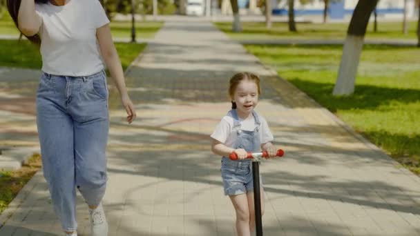 Sağlıklı çocuk şehir caddesi boyunca annesinin gözetiminde scooter kullanıyor. Aile, çocukluk. Mutlu bebek ve anne parkta oynuyorlar. Küçük kız scooter sürmeyi öğreniyor. Doğada aile hafta sonu. — Stok video