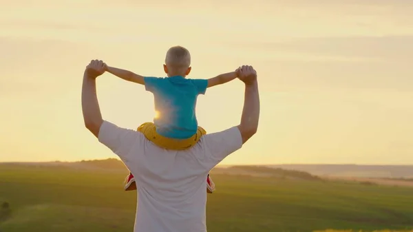 Vader loopt met baby op zijn schouders, heft zijn armen op en vliegt als een vliegtuig. Papa speelt met zijn zoon, gedragen op de schouders van zijn geliefde kind in de zomer op het veld. Gelukkige familie speelt in het park. — Stockfoto