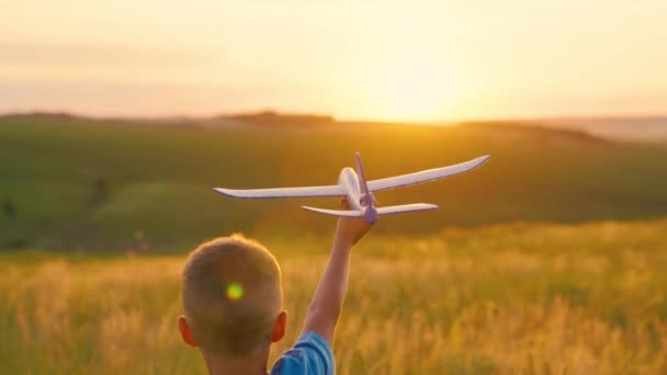 Copil, băiatul aleargă cu un avion de jucărie pe câmpul de vară în raze de apus de soare. Un copil fericit alergând în parc, jucându-se cu avionul de jucărie în natură. O familie fericită. Băiatul visează să zboare. Copilul fără griji se joacă în aer liber — Videoclip de stoc