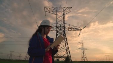 Beyaz kasklı kadın güç mühendisi tabletteki elektrik sensörlerinden elde edilen verileri kullanarak güç hattını kontrol ediyor. Gün batımında yüksek voltajlı elektrik hatları. Dağıtım ve elektrik tedariği. temiz enerji