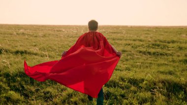 Mutlu çocuk, çocuk süper kahraman, kırmızı pelerinli yeşil alanda koşuyor, pelerin rüzgarda dalgalanıyor. Çocuk oyunları, rüyalar. Ağır çekim. Gençler süper kahraman olmayı hayal eder. Kırmızı pelerin, rüya ifadesi.