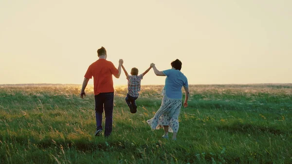 Pappa, son, mamma, lek tillsammans springa, lycklig barn håller händerna på föräldrar, barn hoppar på grönt gräs. Familjevandring i parken på våren vid solnedgången, frisk barndom. Familjehelg på sommaren i naturen — Stockfoto