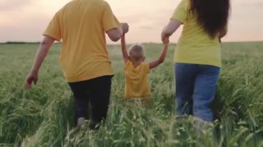 Mutlu aile bahar ve yaz aylarında el ele tutuşarak koşar. Takım çalışması. Aile hafta sonu. Anne, baba ve oğul yeşil buğday tarlasında oynuyorlar, mutlu bir çocuk anne ve babasının elini tutuyor ve zıplıyor..