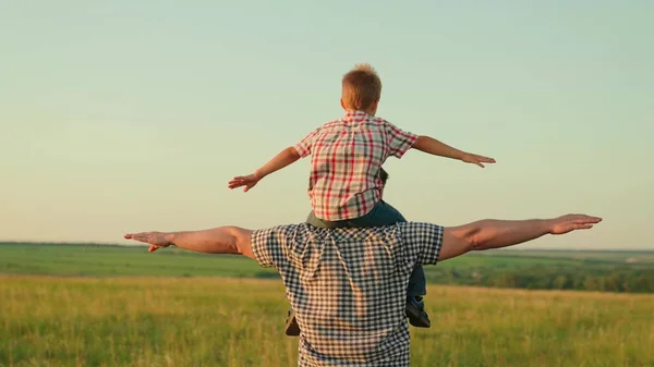 O pai anda com o bebé nos ombros, levanta os braços e voa como um avião. Papai brinca com seu filho, carregado nos ombros de seu filho amado no verão no campo. Família feliz está jogando no parque. — Fotografia de Stock