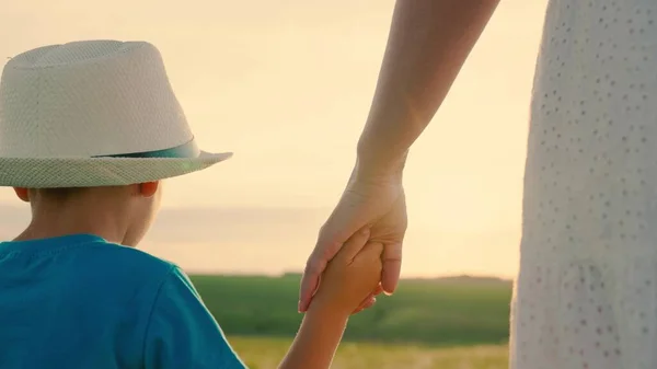 Happy family outing. Mom and son are walking together holding hands in sun. Child in summer hat. People in park. Kid, boy, mom outdoors. Happy family childhood. Parents, cheerful children, dream