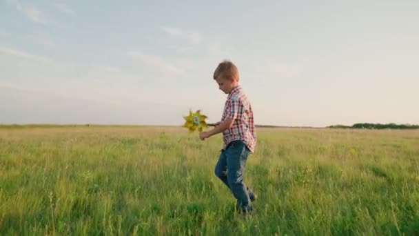 Niño feliz juega con el molinete de juguete al aire libre en el parque de primavera bajo el sol. Infancia, niños. El niño corre con una turbina de viento de juguete en la mano en el campo de verano al atardecer. Vacaciones familiares en la naturaleza. — Vídeo de stock