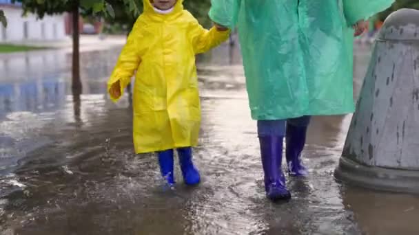 宝贝，他的妈妈穿着雨衣在夏天的公园里散步，孩子们欢天喜地，水从他脚下喷涌而出。小孩跳进了水坑里.快乐的家庭穿着橡胶靴走过水坑. — 图库视频影像