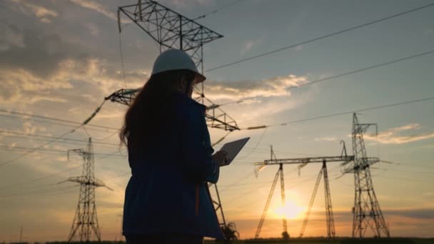 Engineer vrouw power engineer in witte helm controleert power line met behulp van computer tablet. Vrouw Elektriciteitsbedrijf werknemer werkt buiten, diensten hoogspanningsleidingen bij zonsondergang — Stockvideo