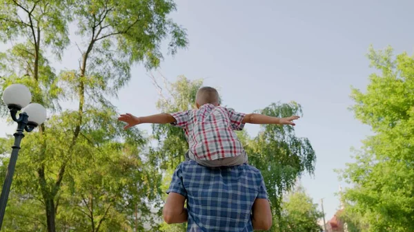 Far går med sin son på axlarna i stadsparken på sommaren. Lycklig familj. På våren leker pappa med barn, barn och far leker tillsammans utomhus. Barn och förälder, friluftsliv, lagarbete. — Stockfoto