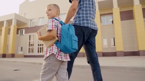 En liten son, skolpojke, går nerför gatan med ryggsäck och håller sin pappa för hand. Förskoleutbildning. Lycklig familj, far och barn går i skolan tillsammans och håller varandra i handen. Pojken går och pluggar. — Stockvideo
