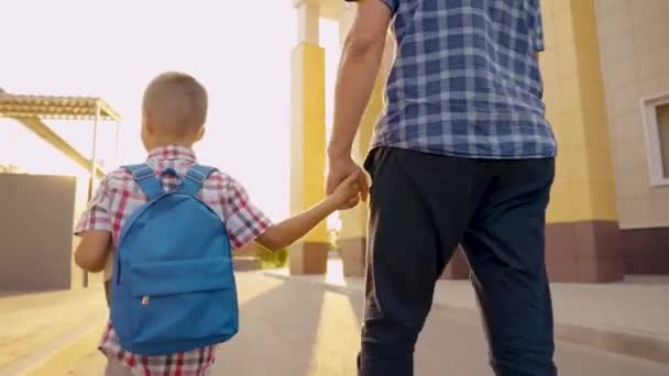 Gelukkige familie, vader en kind gaan samen naar school, hand in hand. Een kleine zoon, schooljongen, loopt door de straat met rugzak en houdt zijn vader met de hand vast. Schoolonderwijs. Baby, jongen gaat studeren. — Stockvideo