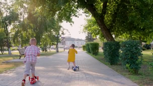 Kız ve erkek açık havada scooter 'a biniyorlar. Sokakta parkta oynayan mutlu şirin çocuklar, scooter 'da denge kurmayı öğreniyorlar. Küçük oğlan ve kız çocuk parkında kaygısız oynuyorlar. Aile yaz tatili — Stok video
