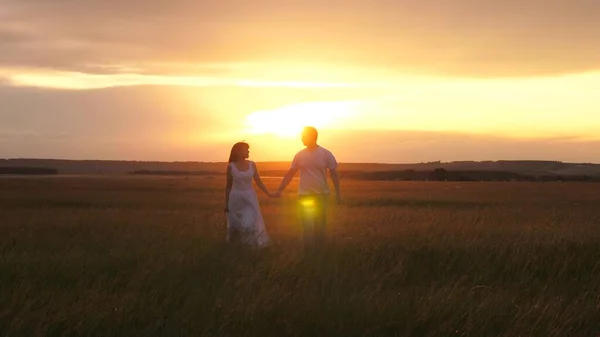 A beautiful girl and a guy are walking hand in hand in field, in sun on the green grass. Silhouette of man and a woman at sunset. A young couple in love is traveling. Romantic date and love in nature. — Stock Photo, Image