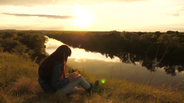 Uno studente legge un libro di testo nel parco all'aperto ai raggi del tramonto. Una giovane viaggiatrice legge il libro ad un punto morto. Turista ragazza con un libro sulla montagna al sole sul fiume. La preghiera in natura. — Video Stock