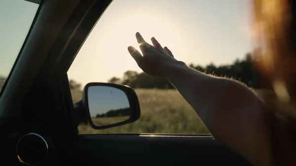 Chica con el pelo largo está sentado en el asiento delantero del coche, estirando su brazo por la ventana y la captura de resplandor de puesta de sol. Mujer libre viaja en coche coge el viento con su mano desde la ventana del coche. Vacaciones —  Fotos de Stock