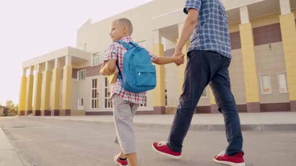 Skolutbildning. Pojken går och pluggar. Lycklig familj, far och barn går i skolan tillsammans och håller varandra i handen. En liten son, skolpojke, går nerför gatan med ryggsäck och håller sin pappa för hand. — Stockvideo