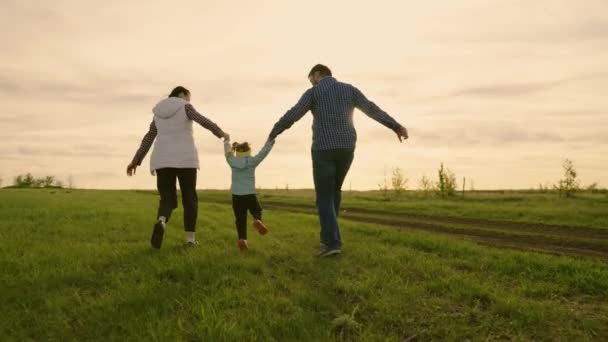 Joyeux famille courir ensemble dans le parc sur l'herbe verte, se tenant la main en été au coucher du soleil. Maman, papa et fille jouent sur le terrain, l'enfant tient la main des parents, saute. Travail d'équipe. Bonne enfance — Video