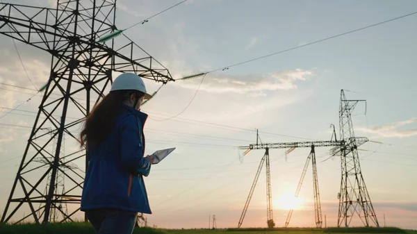 Mujer Electricidad empresa de suministro empleado trabaja al aire libre, servicios de líneas eléctricas de alto voltaje al atardecer. Ingeniera mujer ingeniero de potencia en el casco blanco comprueba la línea de alimentación utilizando la tableta de ordenador —  Fotos de Stock