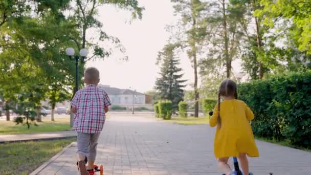 A lány és a fiú együtt robogóznak a szabadban. Boldog aranyos gyerekek játszanak a parkban az utcán, megtanulnak egyensúlyozni a robogón. A kisfiú és a lány gondtalanul játszanak a Childrens Parkban. Családi nyári hétvége — Stock videók