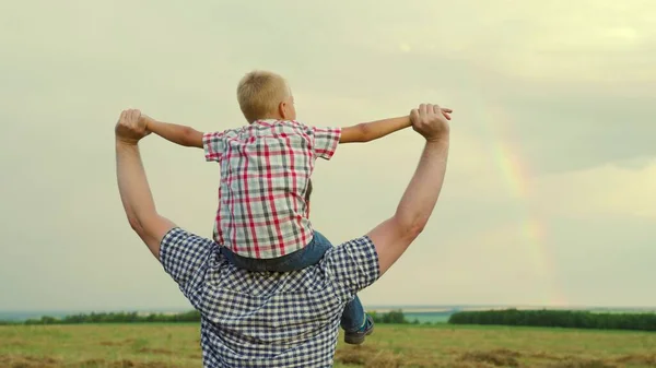 一个快乐的孩子，儿子坐在他父亲的脖子上，扮演飞行员，像超级英雄一样飞翔，梦想着和他的父亲一起飞翔。爸爸和孩子的游戏，幻想。快乐的一家人在天上彩虹前的公园里休息 — 图库照片