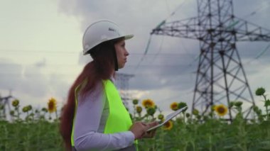 Çevre dostu elektrik enerjisi. Beyaz kasklı bir enerji kadını, tabletteki verileri kullanarak ayçiçeği tarlasında güç hattını kontrol ediyor. Gün batımında yüksek voltajlı elektrik hatları. Elektrik tedariği.