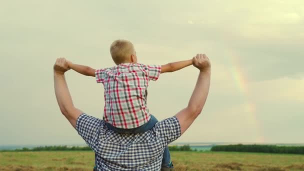 Ett lyckligt barn, en son sitter på sina fäders hals, leker pilot, flyger som en superhjälte, drömmer om att flyga med sin pappa. Pappa och barn leker, fantiserar. Lycklig familj vilar i parken framför regnbågen på himlen — Stockvideo