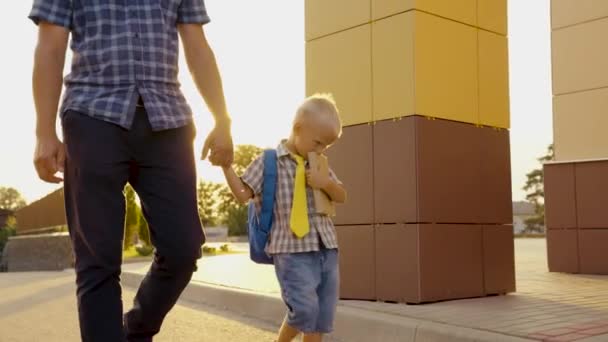 Dad is taking his little son with textbook to school. Schoolboy boy walking down street with backpack, holding his dad by hand. Preschool education. Happy family, father, child go to school together — Stock Video