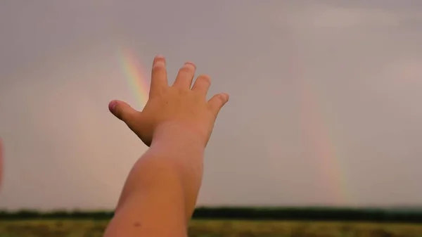 Happy child, boy stretches out his hand to the rainbow against the blue sky. The child dreams of touching a multi-colored rainbow in the sky. Family walks in the park after rain. Happy family concept