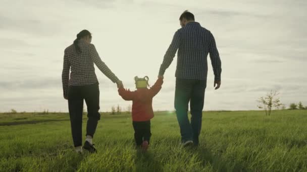 Familienwochenende. Mama, Papa und Tochter gehen, Händchen haltend, Kind hüpft auf dem grünen Gras. Fröhliche Familienwanderung im Park im Frühling bei Sonnenuntergang. Glückliche gesunde Kindheit. — Stockvideo