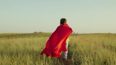 Çocuk oyunları ve rüyalar. Mutlu süper kahraman kız, yeşil sahada kırmızı pelerinle koşuyor, pelerin rüzgarda dalgalanıyor. Gençler süper kahraman olmayı hayal eder. Kırmızı pelerinli genç kız, rüya ifadesi..