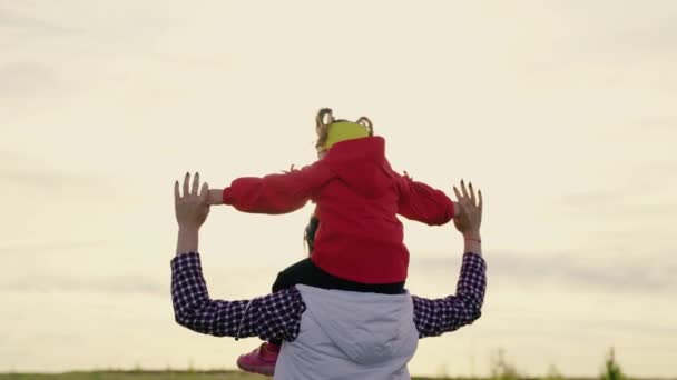 Mama bawi się z córką i nosi swoje ukochane dziecko na ramionach w słońcu. Dziecko i rodzice bawią się razem. Szczęśliwa rodzina w parku. Matka chodzi z dzieckiem na ramionach w promieniach zachodzącego słońca. — Wideo stockowe