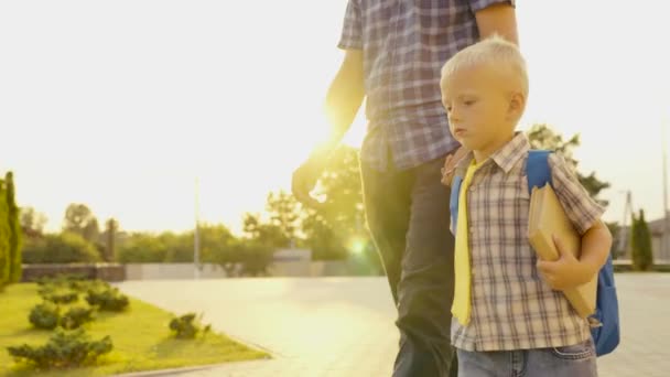 Lycklig familj, far, barn går i skolan tillsammans. Pappa tar sin son med sig till skolan. Skolpojke pojke gå ner gatan med ryggsäck, hålla sin pappa för hand. Förskoleutbildning. — Stockvideo