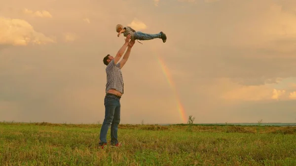 Dad throws his happy son into sky in park in front of a rainbow. Father, child play, laugh and rejoice together. Happy family trip. The baby is in arms of a parent. Dad is off. Happy family concept — Zdjęcie stockowe