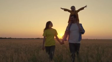 Mutlu aile gün batımında buğday tarlasında yürüyor. Bir çiftçi ailesi. Çocuk babasının omuzlarında, annesinin yanında, parkta el ele tutuşuyor. Ebeveynler, sevgili bebeğim, güneş ışınları altında mutlu bir şekilde yürüyorlar..