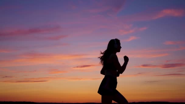 Silhouette of free young woman runs in summer in park at sunset, listens to music on headphones. Training jogging. Healthy jogging and outdoor exercise. Listen to music without Internet, play sports — Stock Video