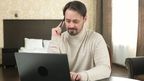 Freelance worker working with modern laptop and cell phone in room. Male professional works using a computer and cell phone in office, at home. Businessman, working on laptop, talking on a smartphone. — ストック写真