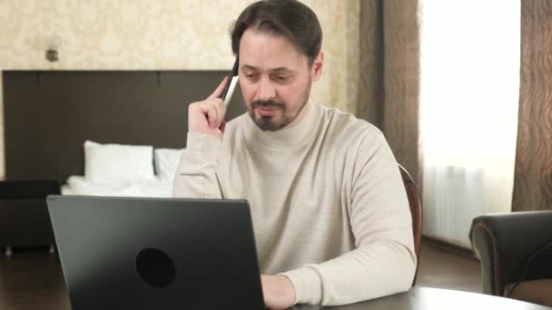 Freelance worker working with modern laptop and cell phone in room. Male professional works using a computer and cell phone in office, at home. Businessman, working on laptop, talking on a smartphone. — Vídeos de Stock