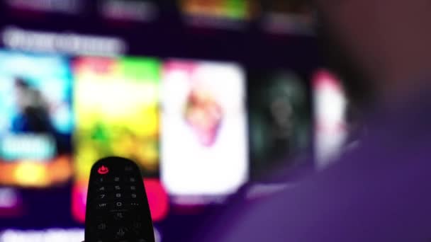 Een man kijkt smart TV en gebruikt zwarte afstandsbediening. Mans hand selecteert internet tv-kanalen met afstandsbediening, close-up. Persoon bestuurt TV met behulp van een moderne afstandsbediening. wazige tv scroll pagina 's — Stockvideo