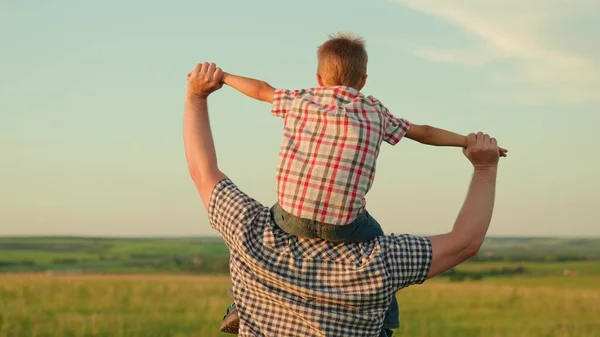 Ojciec chodzi z dzieckiem na ramionach, podnosząc ręce i latając jak samolot. Tata bawi się z synem, nosi go na ramionach ukochanego dziecka latem na polu. Szczęśliwa rodzina bawi się w parku. — Zdjęcie stockowe