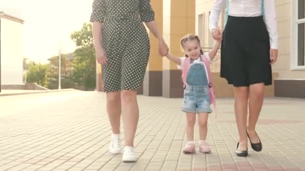 En lycklig familj, mor och barn går i skolan och håller varandra i hand. Glada barn hoppar. Skolutbildning. En liten skolflicka dotter går med ryggsäck och håller sin mor och syster för hand. — Stockvideo